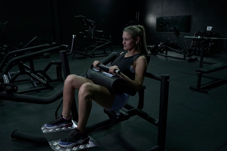 A woman performs leg exercises on a gym machine in a dimly lit environment.
