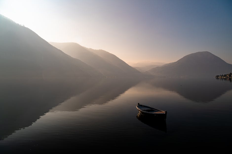Calm water with a lone boat at sunrise, surrounded by misty mountains.