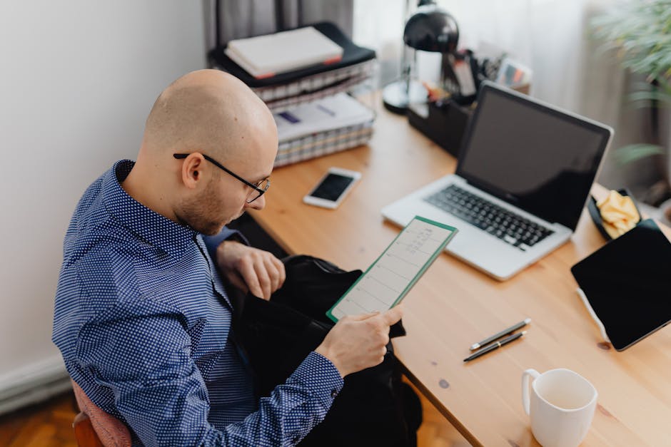 Bald man in blue shirt working at desk with laptop and documents in an office setting.